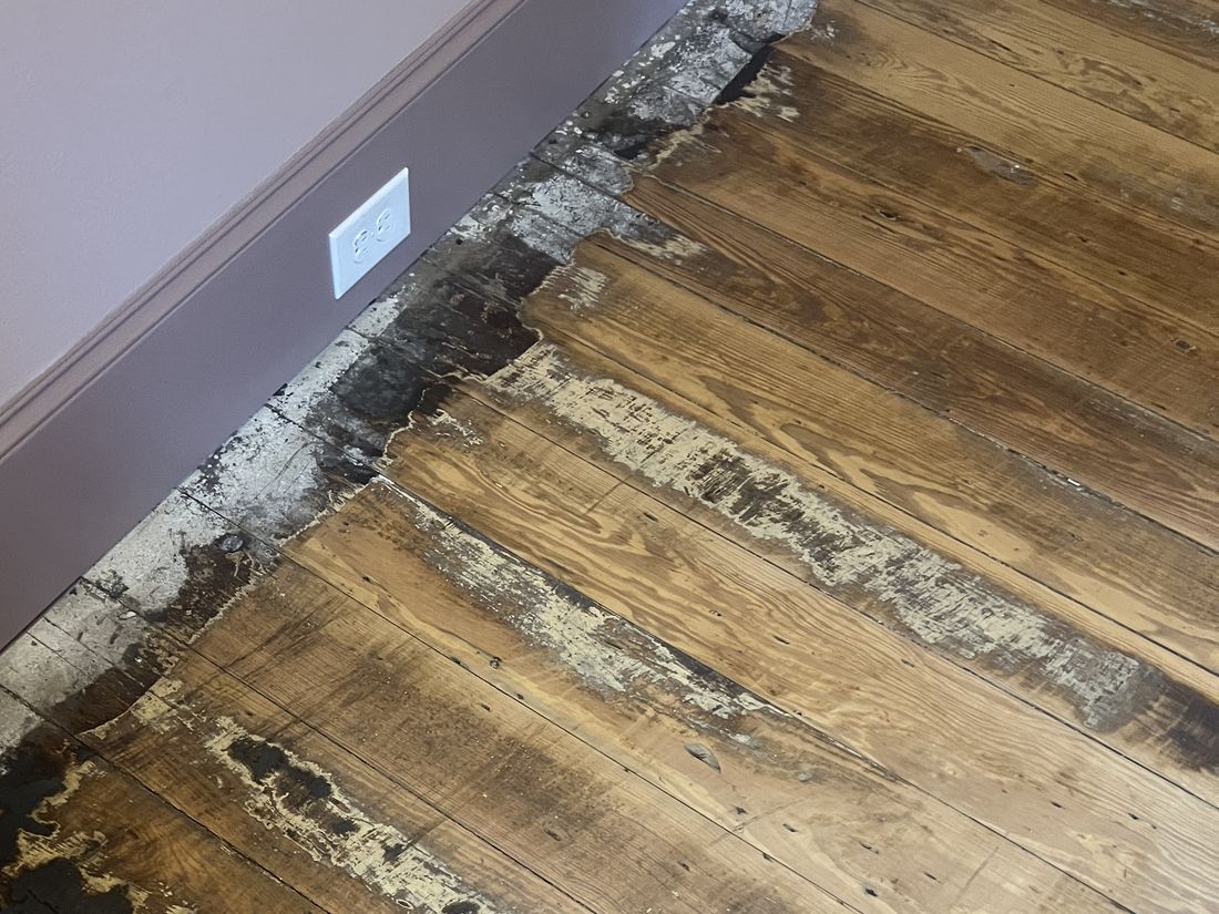 Old floor with rotted boards, paint residue and gaps along the baseboard, before repair